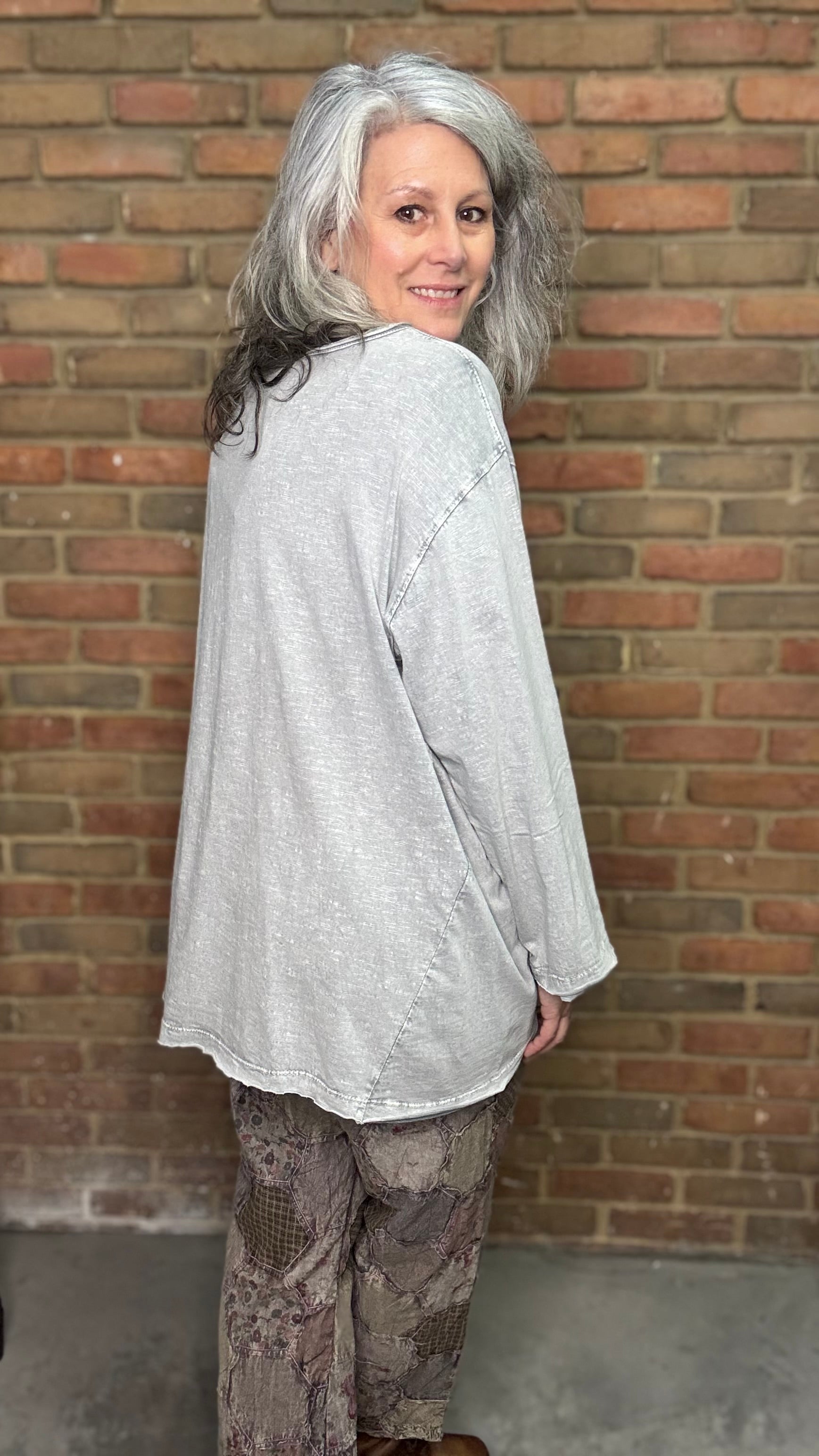 Woman wearing a light gray long-sleeve top and patterned pants standing against a brick wall.