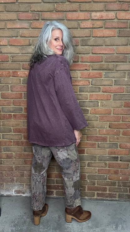 Person wearing a purple jacket and patterned pants standing against a brick wall.