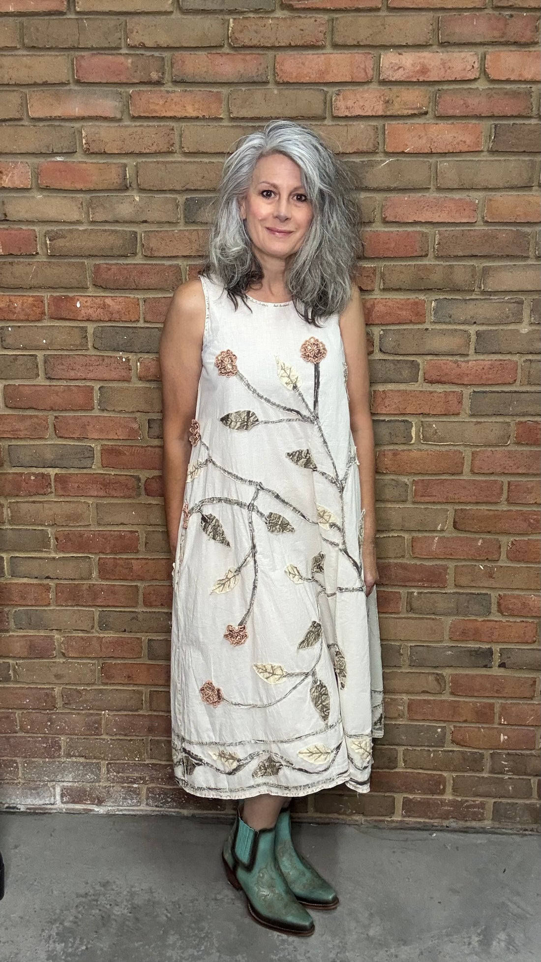 Woman in a white dress with floral patterns standing against a brick wall.