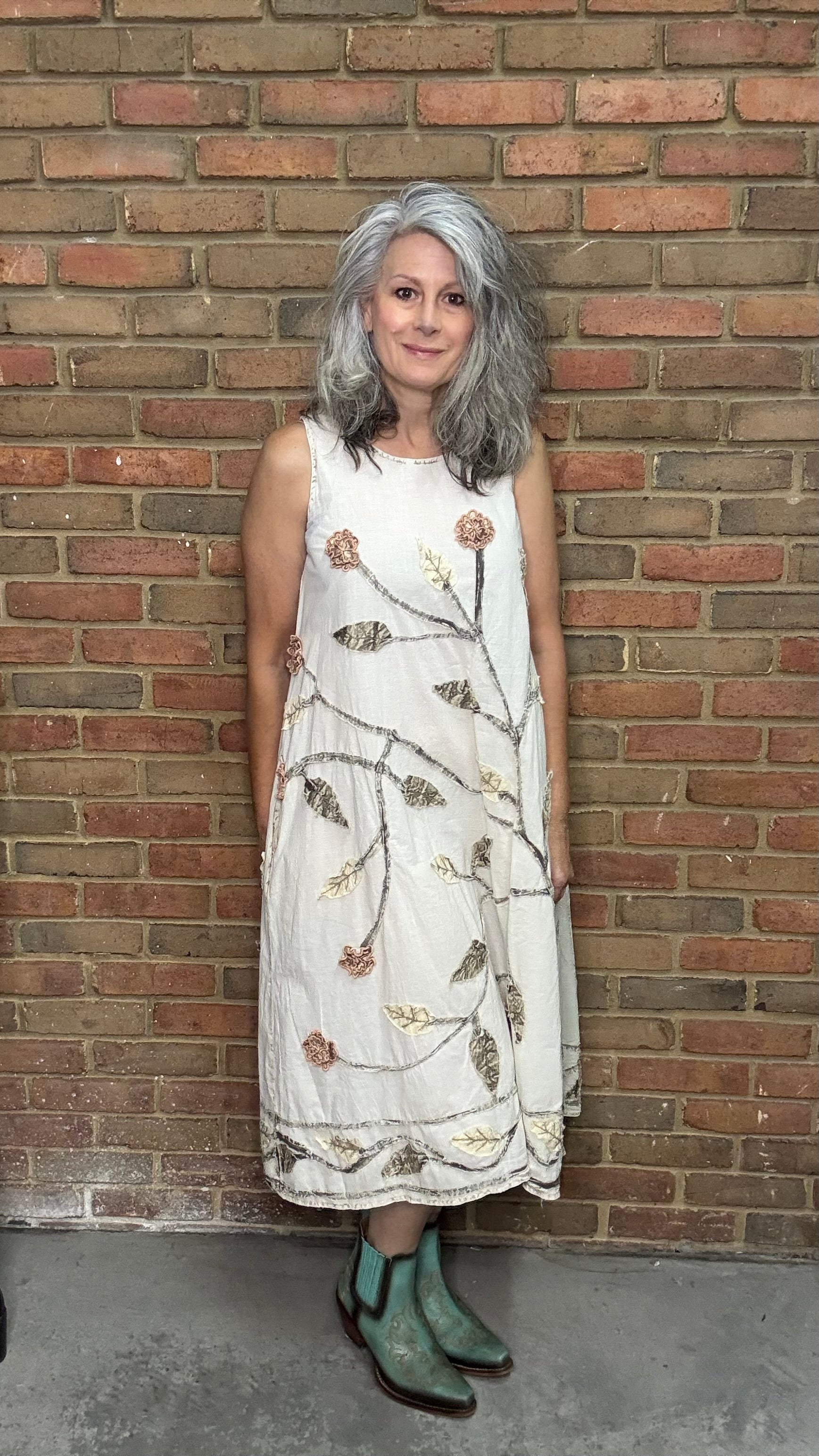 Woman in a white dress with floral patterns standing against a brick wall.