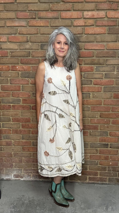 Woman in a white dress with floral patterns standing against a brick wall.