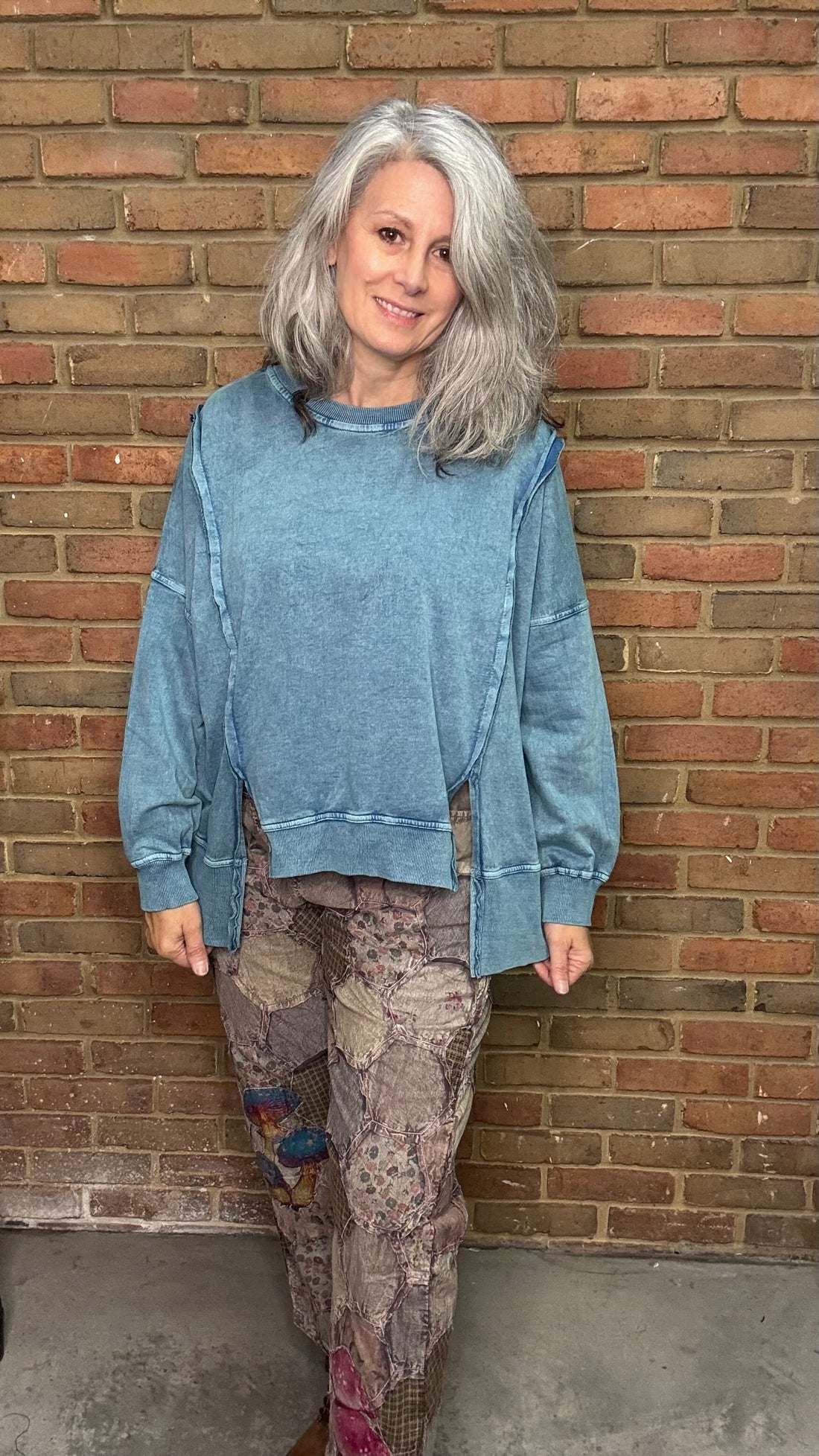 Person wearing a blue sweatshirt and patterned pants standing against a brick wall.