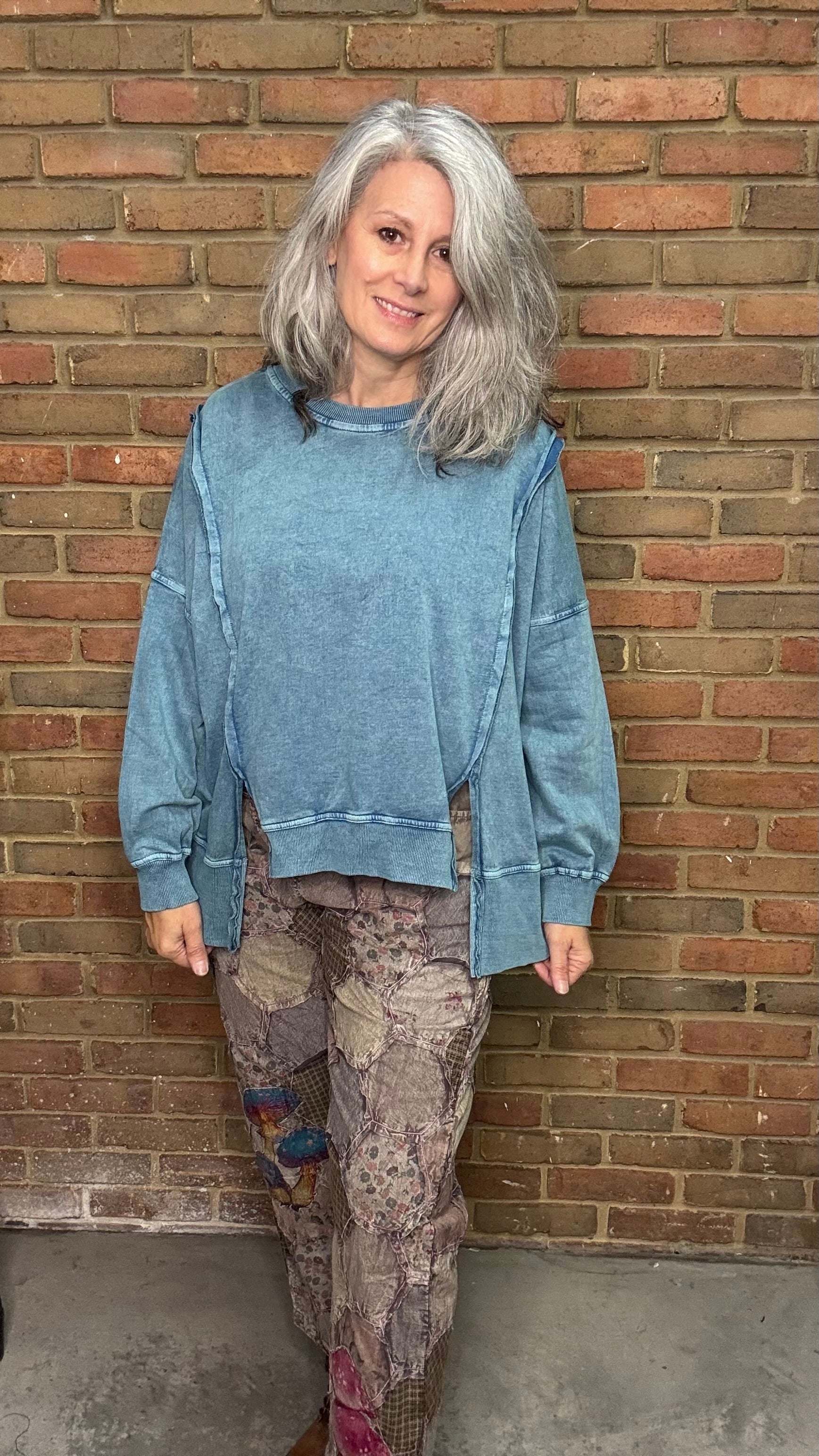 Person wearing a blue sweatshirt and patterned pants standing against a brick wall.