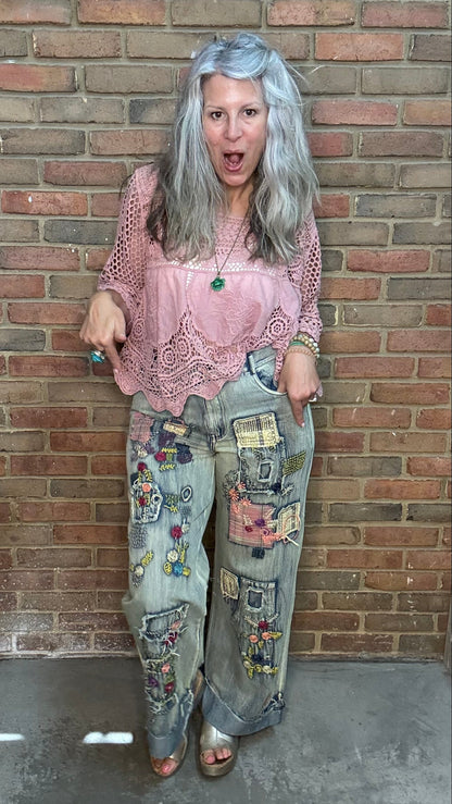 A woman with long gray hair stands against a brick wall wearing a pink crochet top and heavily decorated light-wash jeans covered in colorful patches and patches. She is wearing brown shoes.