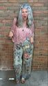 A woman with long gray hair stands against a brick wall wearing a pink crochet top and heavily decorated light-wash jeans covered in colorful patches and patches. She is wearing brown shoes.