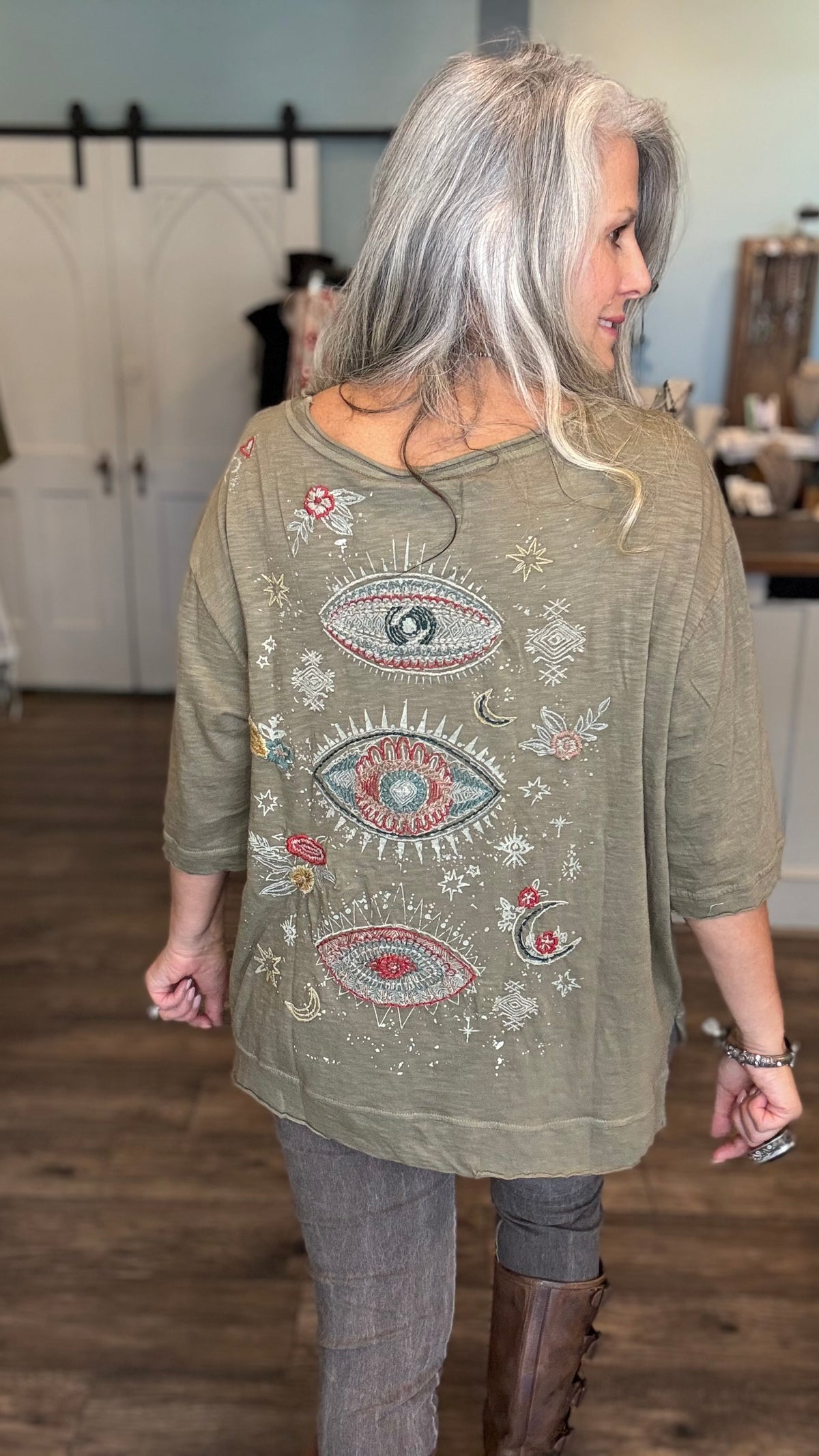 A woman with long silver-gray hair is shown from the back wearing a loose olive green top with colorful embroidered eye and floral motifs, paired with gray pants and brown boots in a boutique setting.