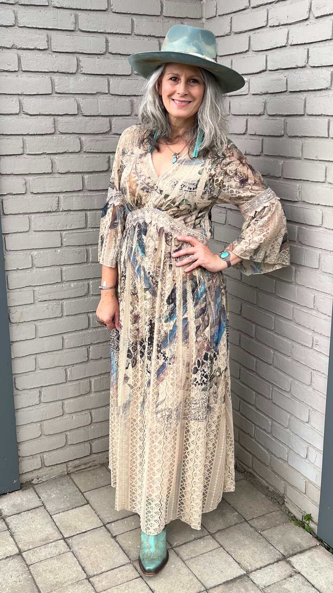 Woman in a floral dress and hat standing against a brick wall.