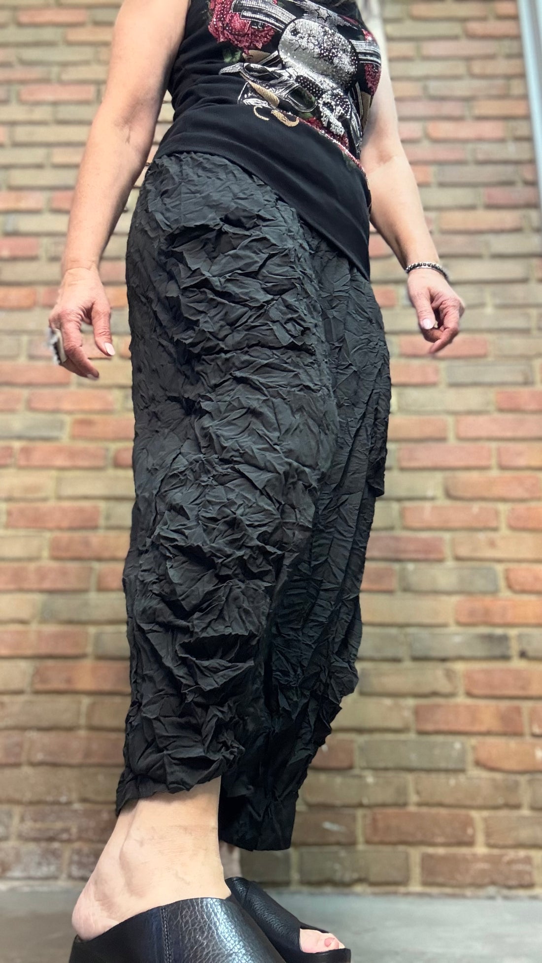 Person wearing Black Crinkle Side Pocket Pant and a patterned top against a brick wall.