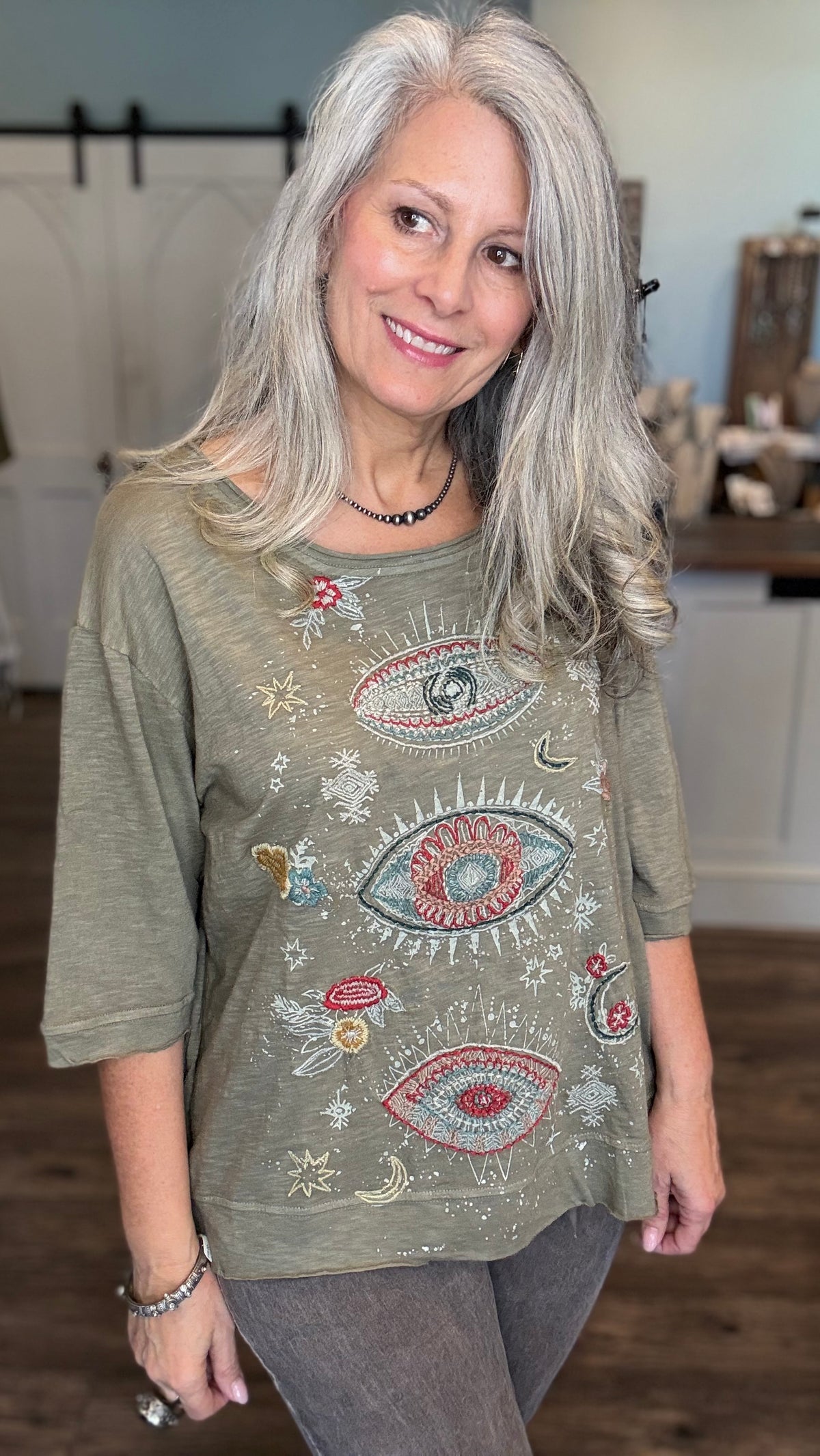 A smiling woman with long gray hair wearing a sage green short-sleeve top with colorful embroidered eyes, stars, and floral details, paired with gray pants in an indoor setting.