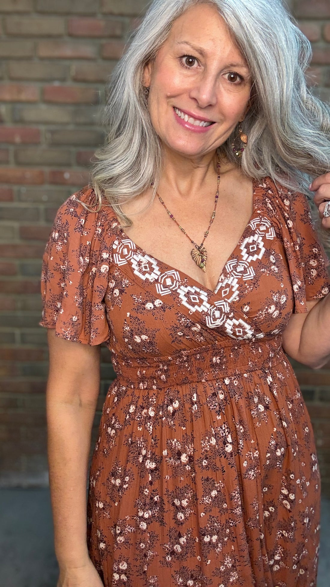 A woman with gray hair stands against a brick wall wearing a rust-brown bohemian-style midi dress with white floral prints, the "Short Sleeve Embroidered Midi Dress" and brown cowboy boots.