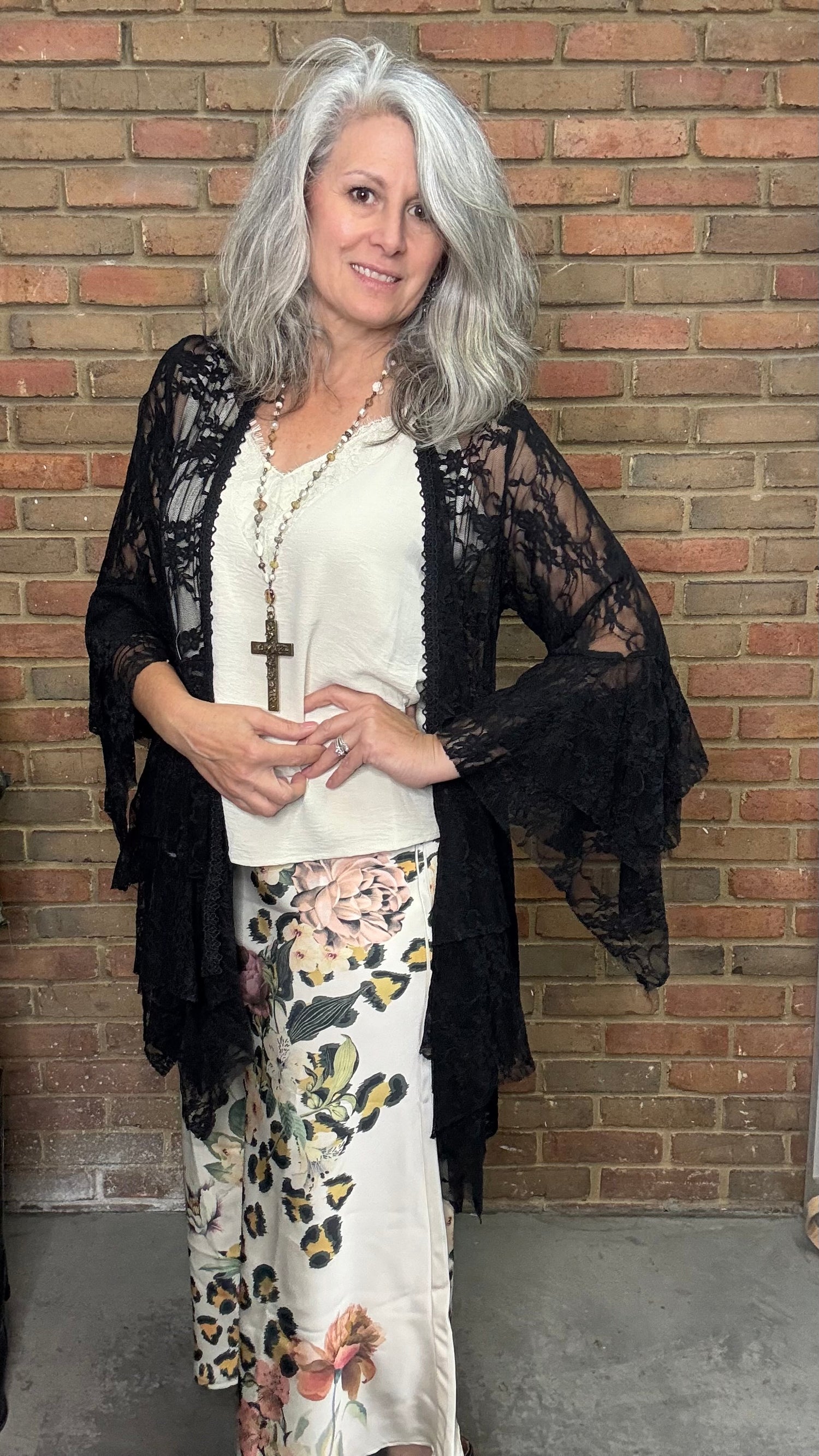 Woman wearing a black lace kimono over a white top and floral pants against a brick wall.
