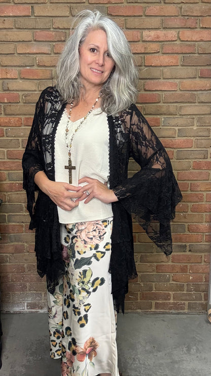 Woman wearing a black lace kimono over a white top and floral pants against a brick wall.