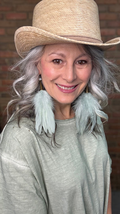 Woman wearing a straw hat with Sage Feather Earrings and a gray top against a brick wall.