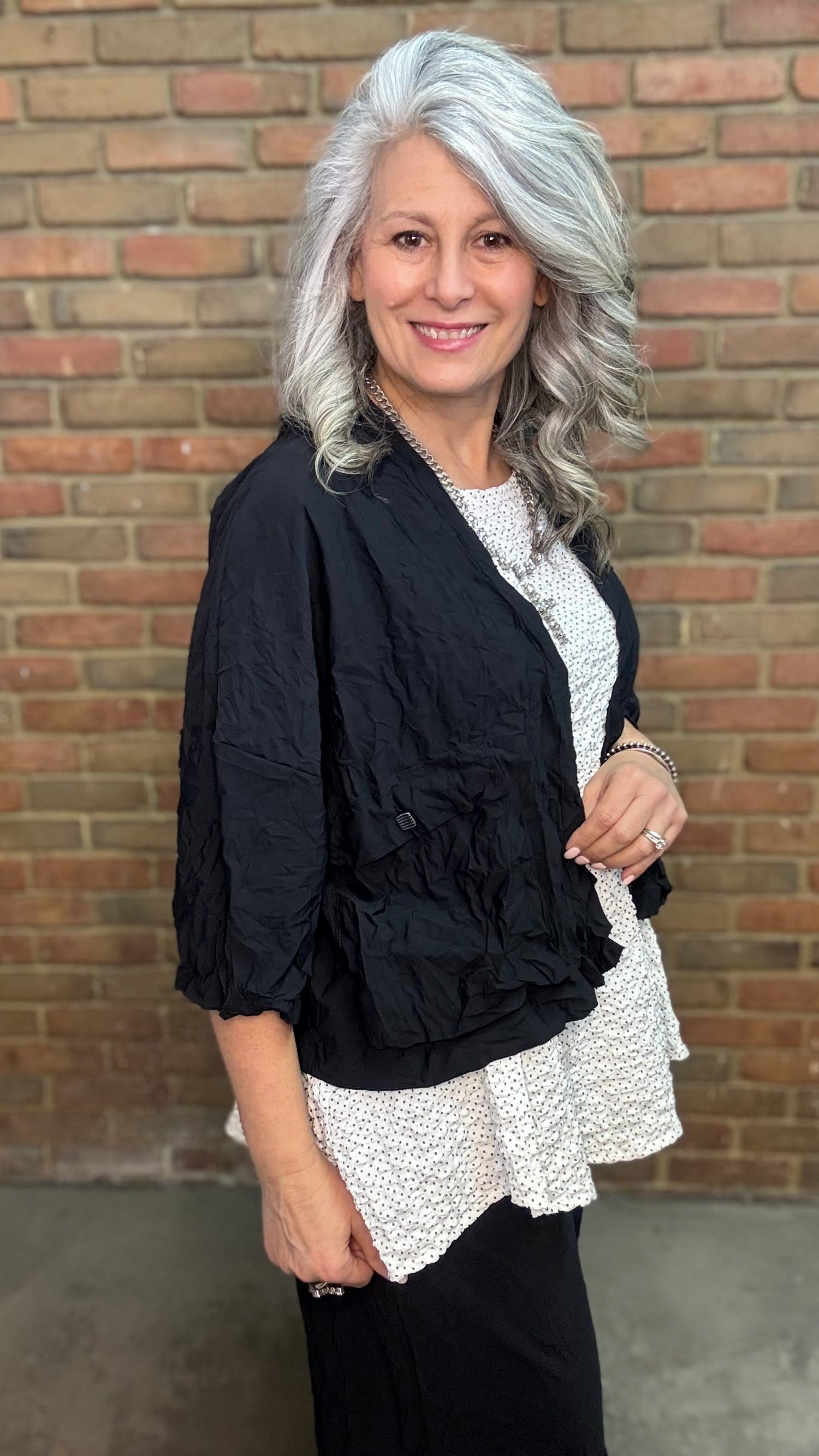 Woman standing against a brick wall wearing Francesca Jacket - Black, white top, and black pants.