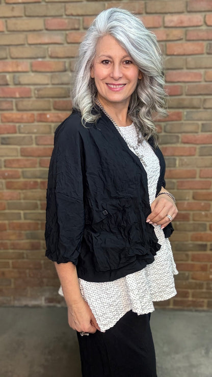 Woman standing against a brick wall wearing Francesca Jacket - Black, white top, and black pants.