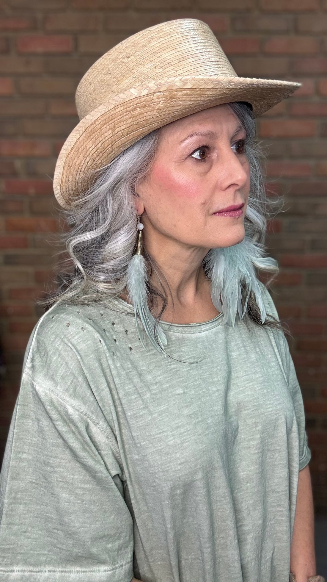 Woman wearing a straw hat with Sage Feather Earrings and a gray top against a brick wall.
