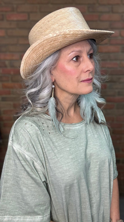Woman wearing a straw hat with Sage Feather Earrings and a gray top against a brick wall.