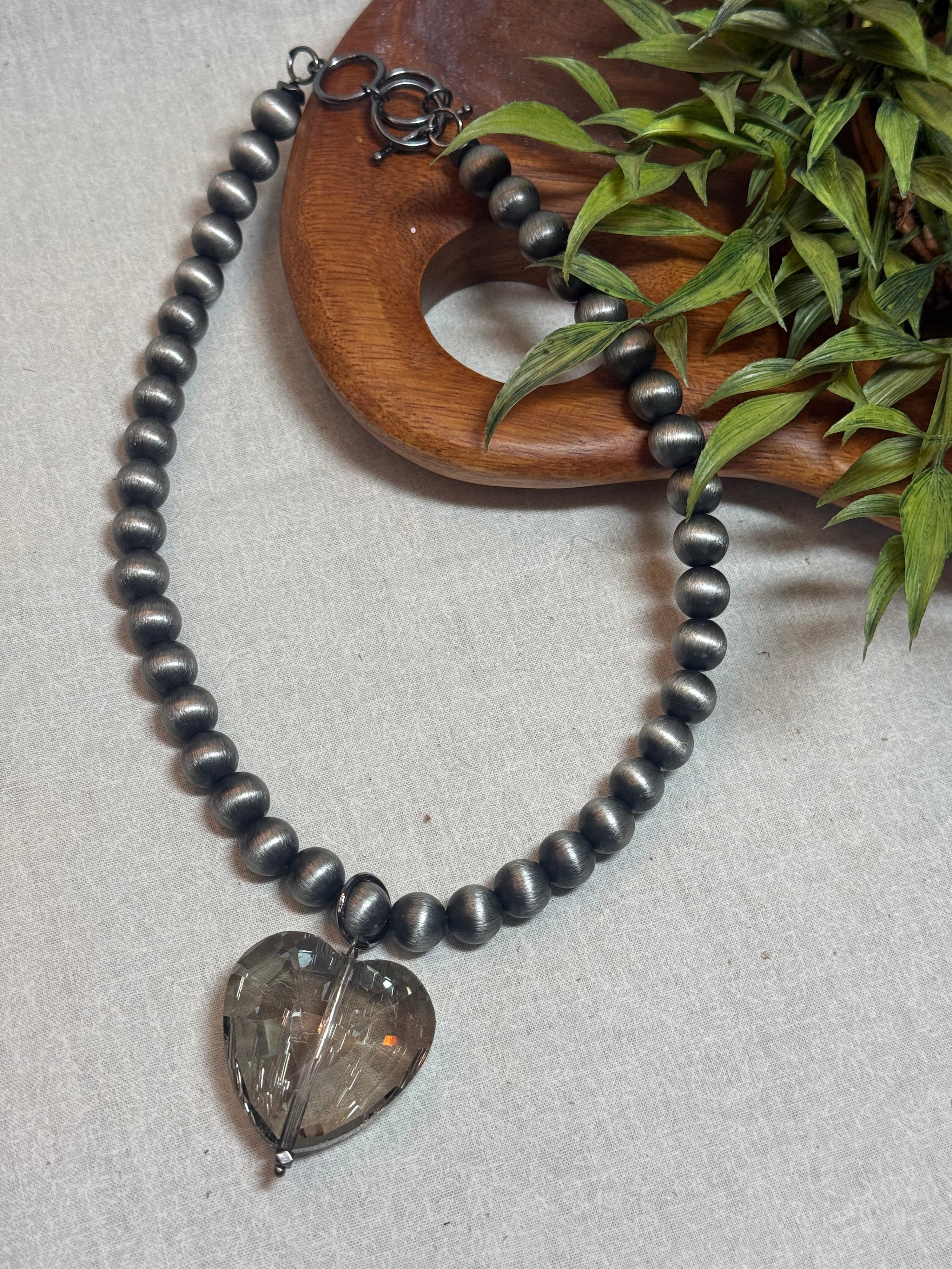 A dark gray beaded necklace with a heart-shaped smoky crystal pendant, styled on a light beige textured surface next to a wooden tray and green leaves.