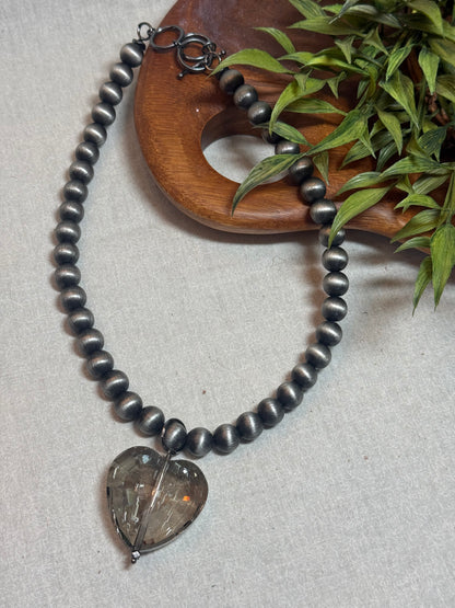 A dark gray beaded necklace with a heart-shaped smoky crystal pendant, styled on a light beige textured surface next to a wooden tray and green leaves.