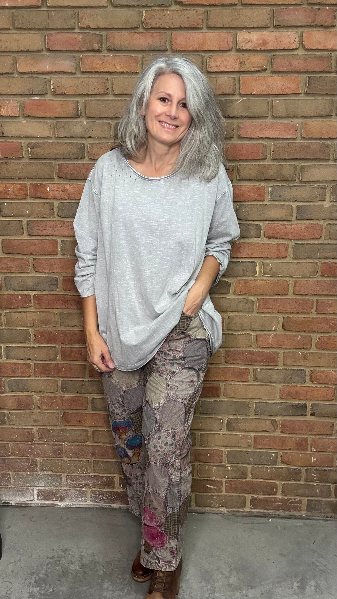Woman leaning against a brick wall wearing a gray sweater and patterned pants.