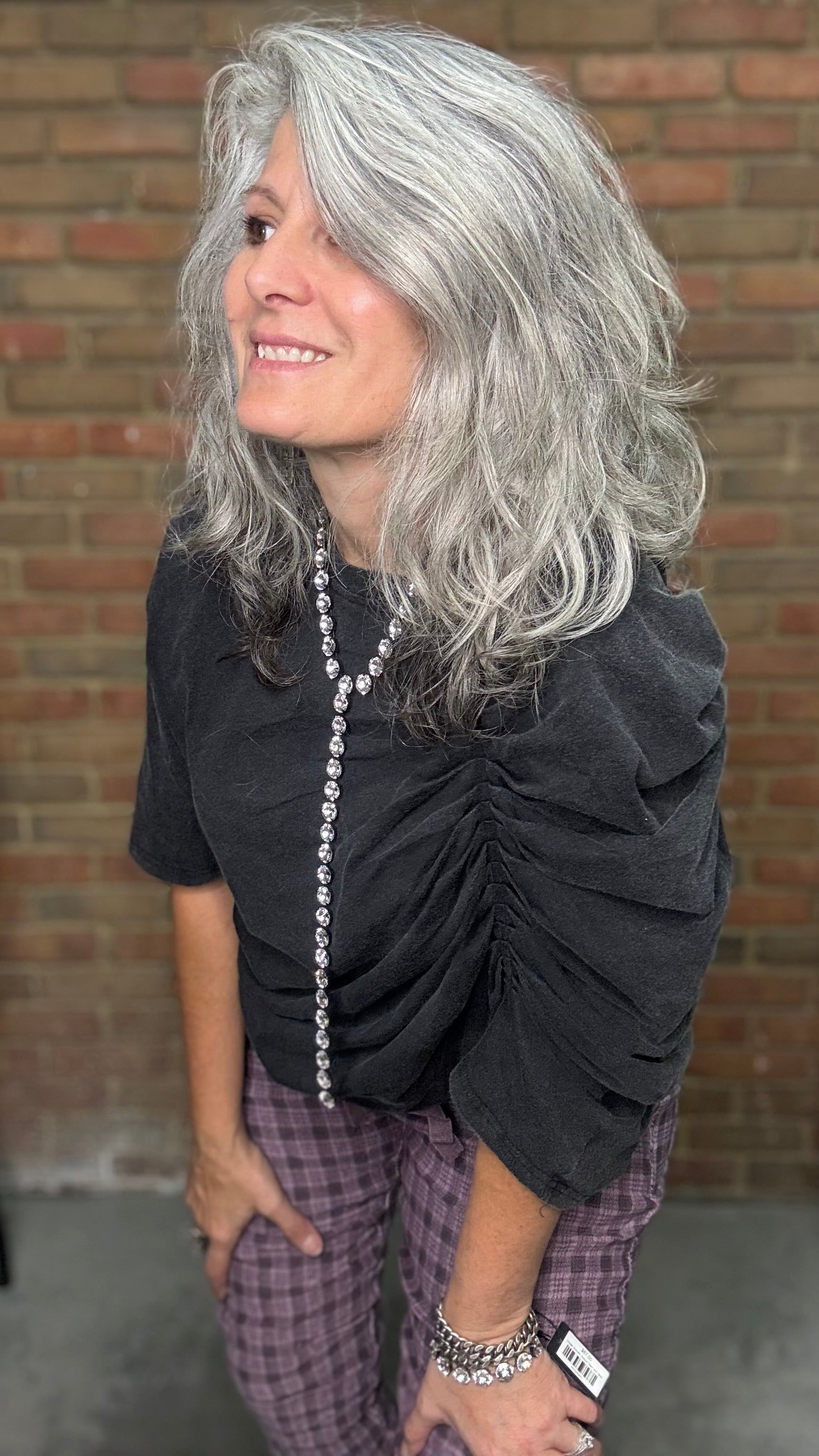 Person with gray hair wearing a black top and plaid pants against a brick wall.