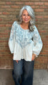 A woman with gray hair wearing a white long-sleeve blouse with a blue floral pattern and dark blue jeans, standing in front of a brick wall.