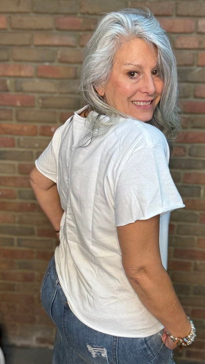 Woman with gray hair wearing a white t-shirt and blue jeans standing against a brick wall.