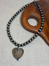 A beaded necklace with dark gray metallic round beads and a heart-shaped smoky glass pendant, displayed on a light gray textured surface next to a wooden prop.