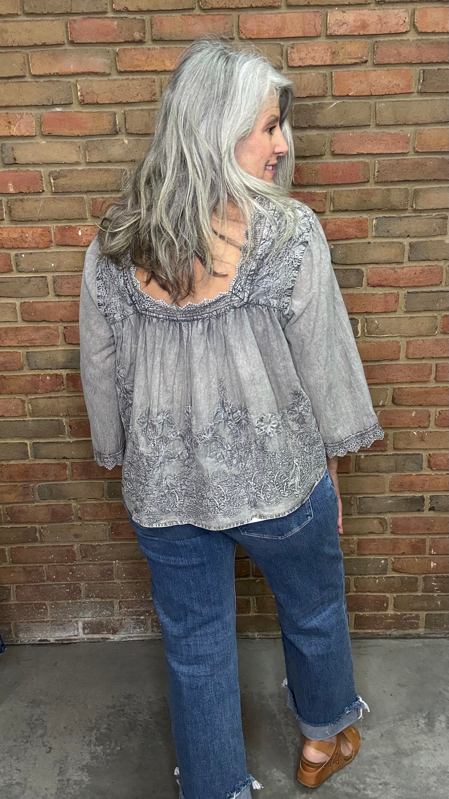 Woman with gray hair wearing the Vintage Washed Floral Lace Top a  light gray blouse and blue jeans standing against a brick wall.