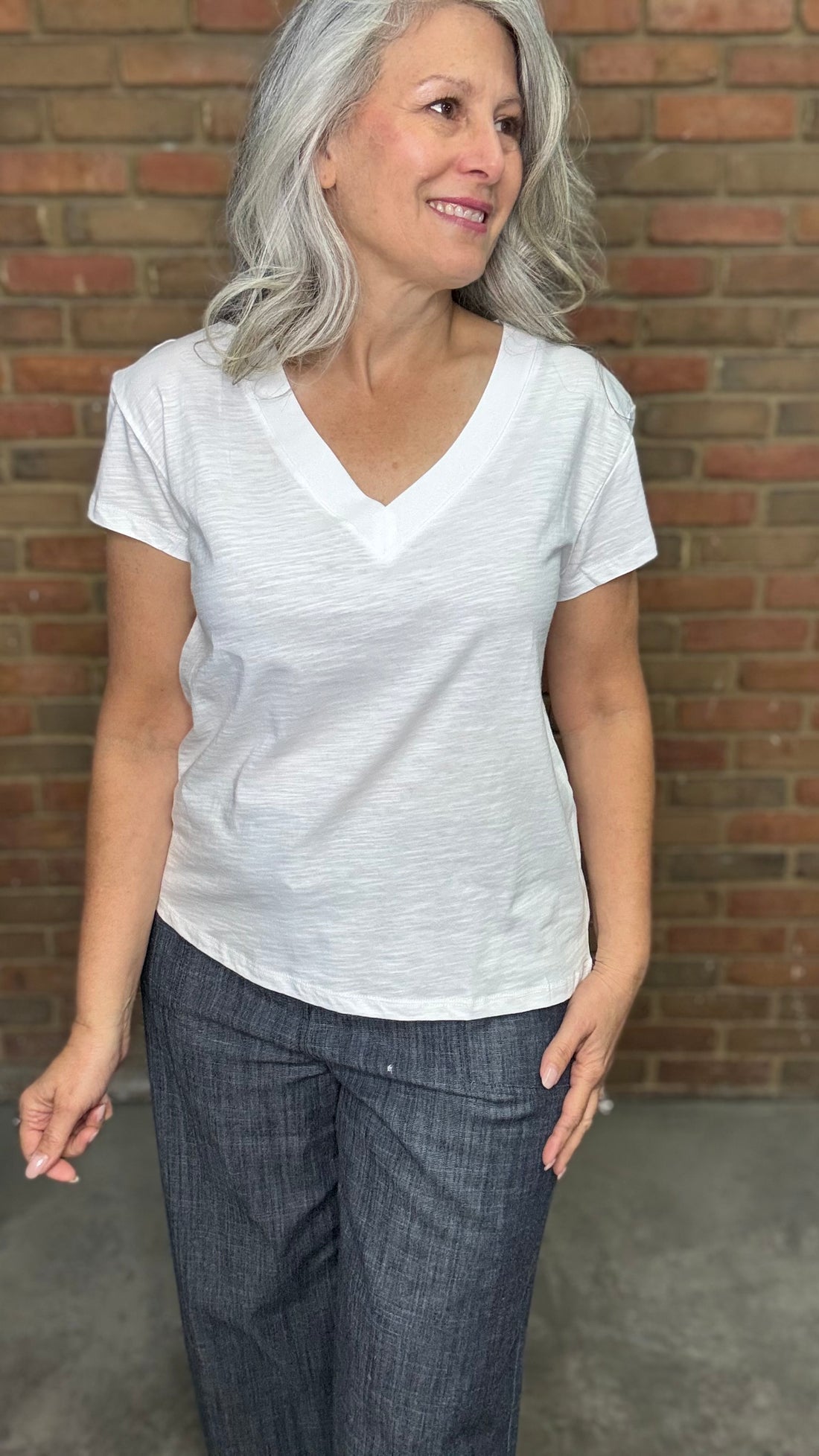 Woman wearing White Drop Shoulder V-Neck Top and Indigo Linen Button Front Pants standing against a brick wall.