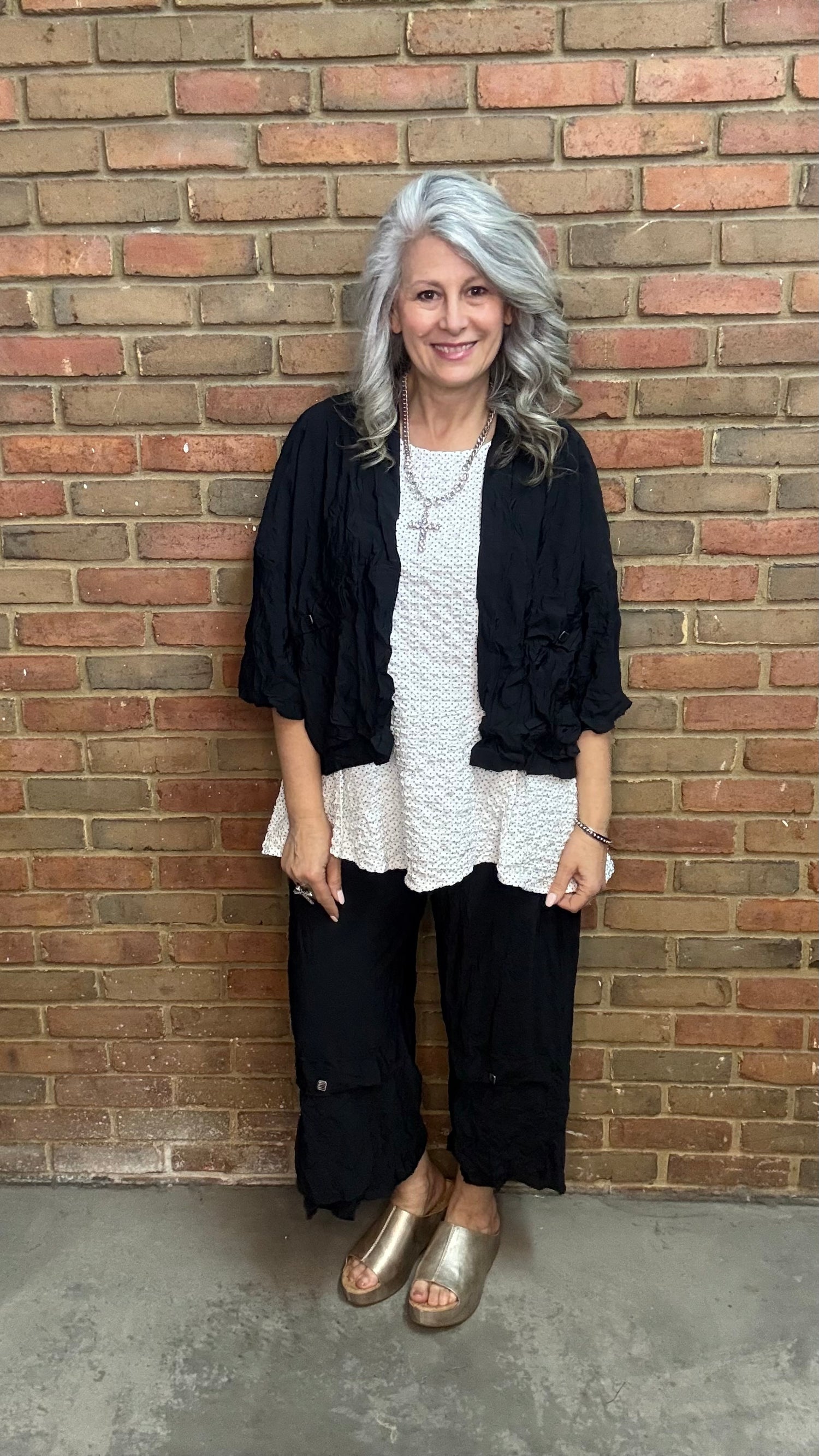 Woman standing against a brick wall wearing Francesca Jacket - Black, white top, and black pants.