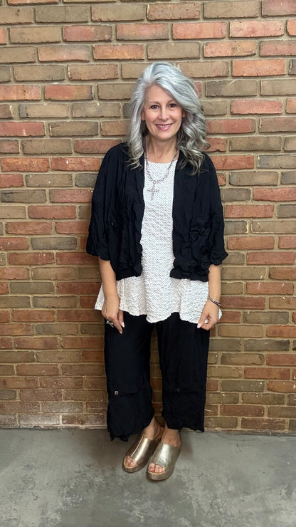 Woman standing against a brick wall wearing Francesca Jacket - Black, white top, and black pants.