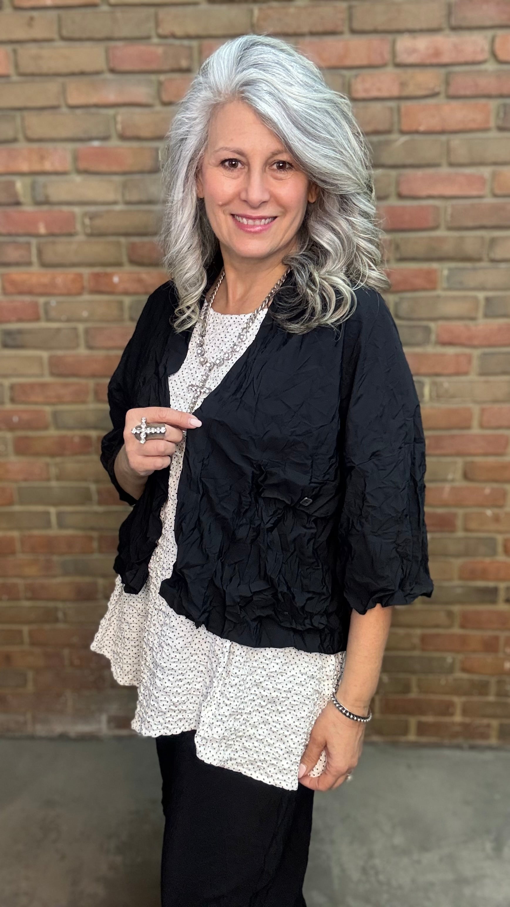 Woman standing against a brick wall wearing Francesca Jacket - Black, white top, and black pants.