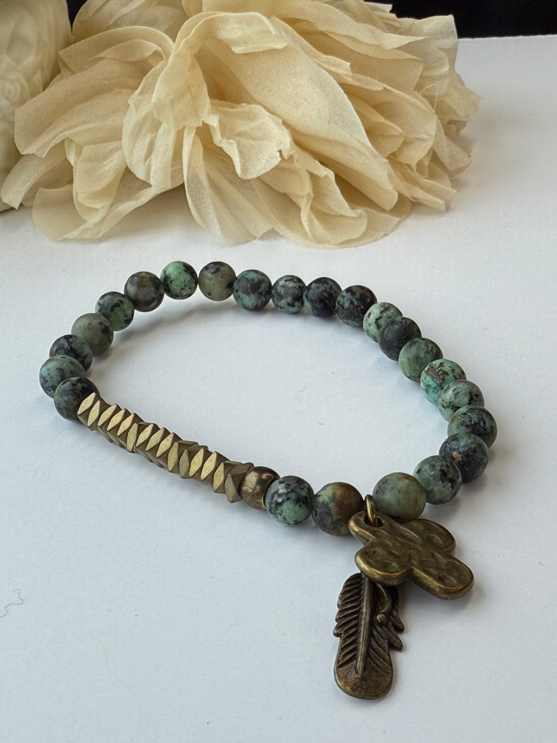 Bracelet with green beads and a bronze feather charm next to a beige flower on a white background