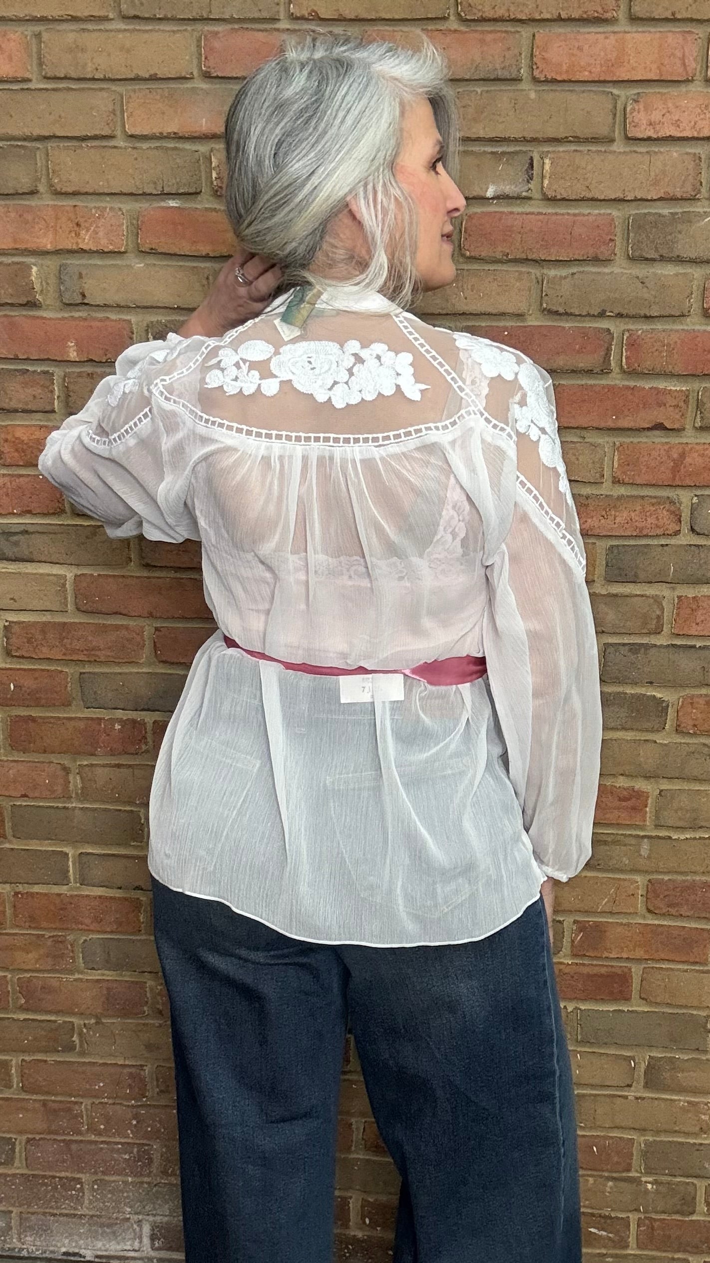 Person wearing a sheer white blouse with floral details and dark blue jeans against a brick wall.