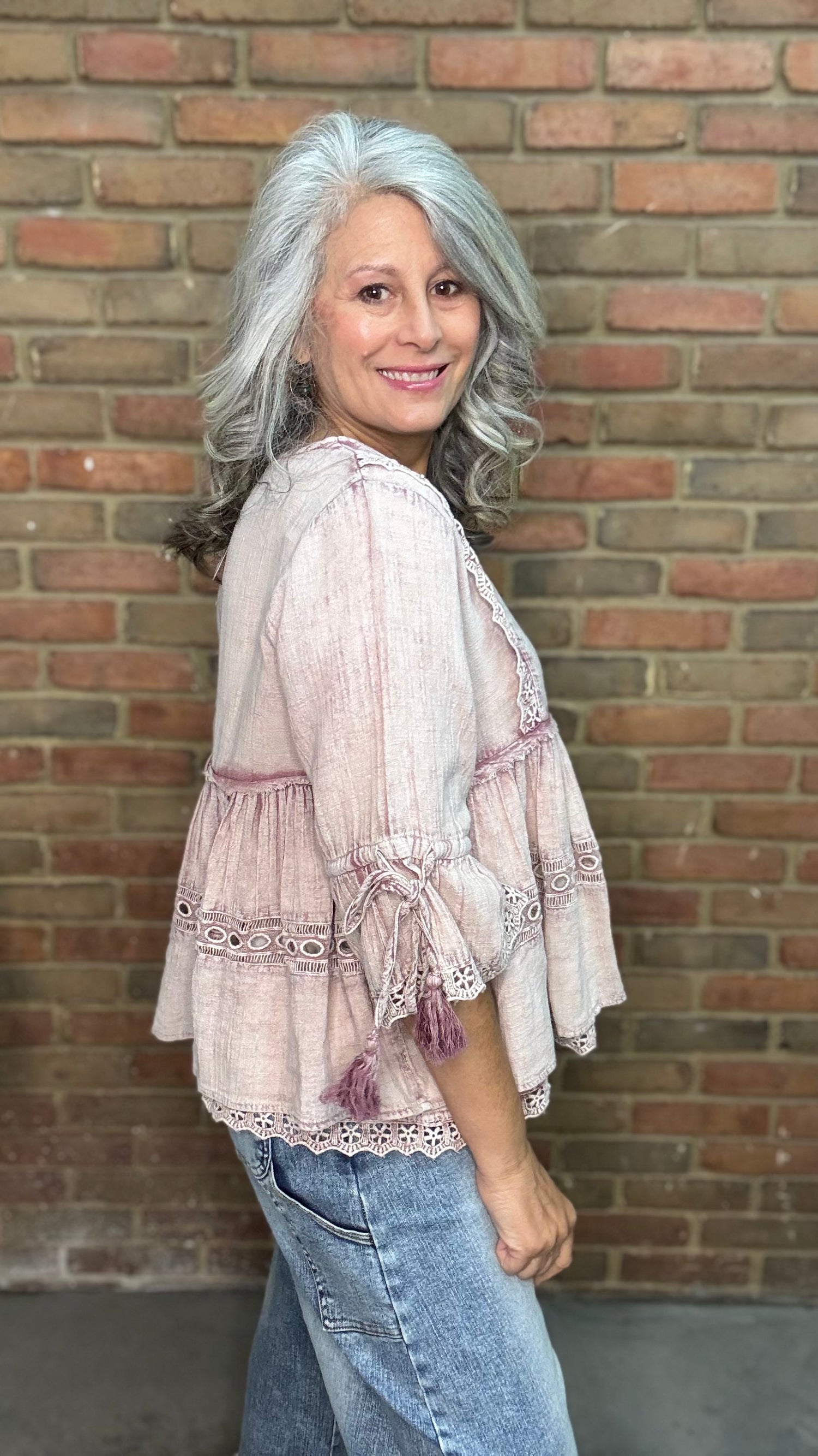 Woman wearing the Washed Pink Cotton Gauze Top, a light pink blouse, and blue jeans standing against a brick wall.
