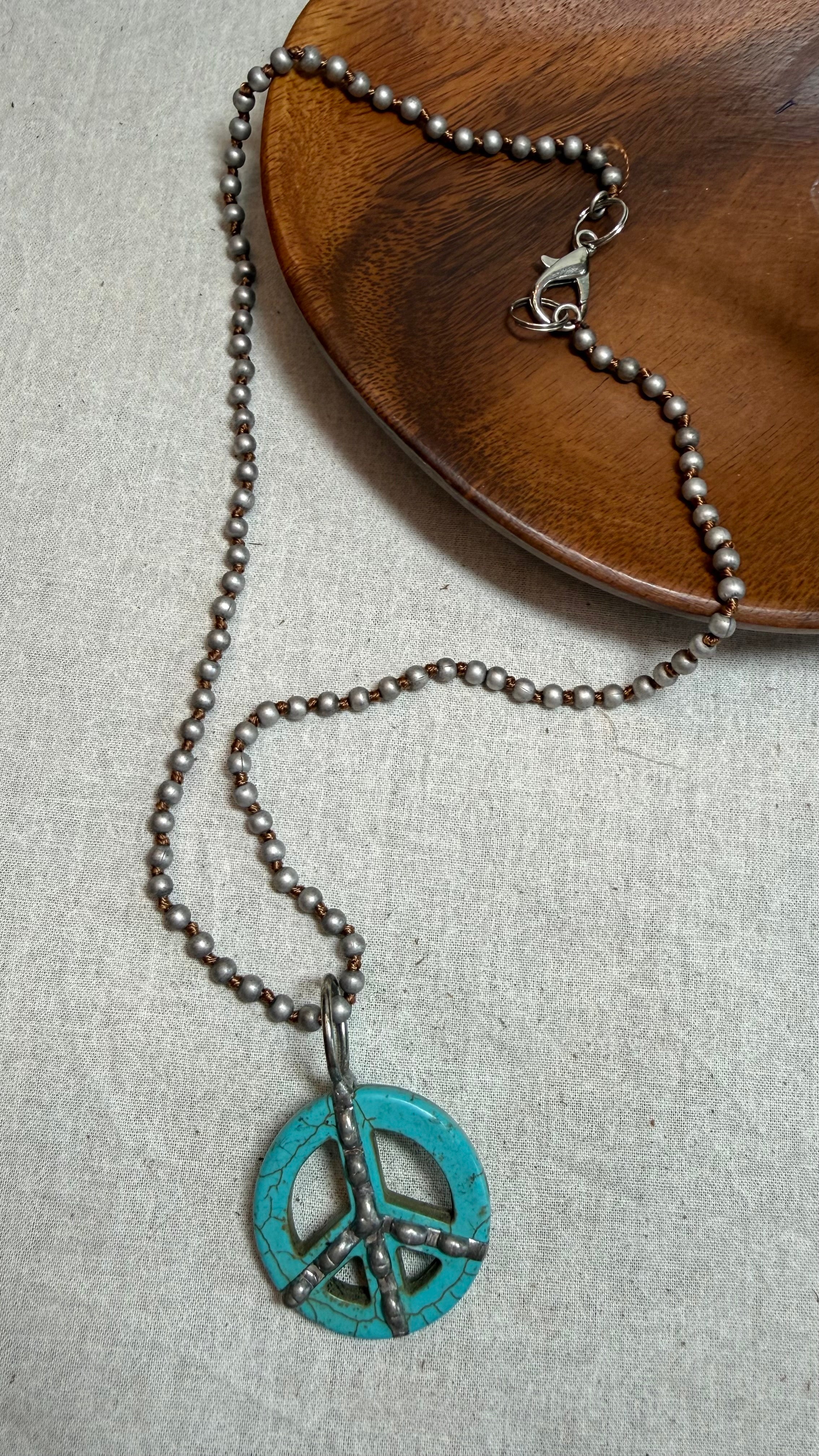 A silver-toned beaded necklace featuring a turquoise peace sign pendant with oxidized black details, displayed on a textured light gray surface with a wooden plate in the background.
