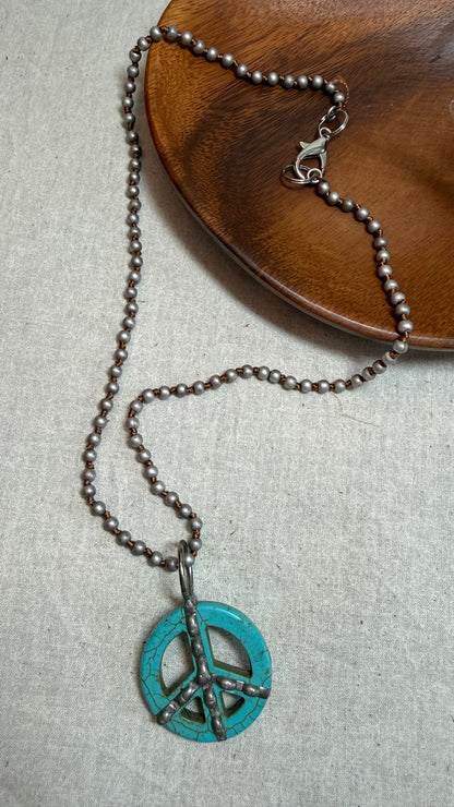 A silver-toned beaded necklace featuring a turquoise peace sign pendant with oxidized black details, displayed on a textured light gray surface with a wooden plate in the background.