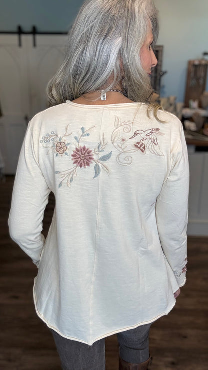 A woman with long silver-gray hair is shown from the back wearing a cream-colored long-sleeve top with delicate floral and bird embroidery on the upper back, paired with gray pants in an indoor setting.
