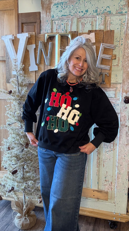 Woman wearing a black sweater with colorful text, standing in front of a Christmas tree and wooden wall.