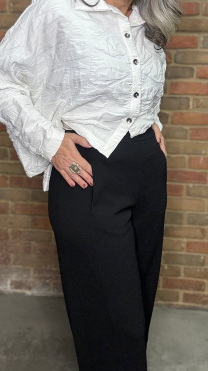 Person wearing a white blouse and black pants against a brick wall.