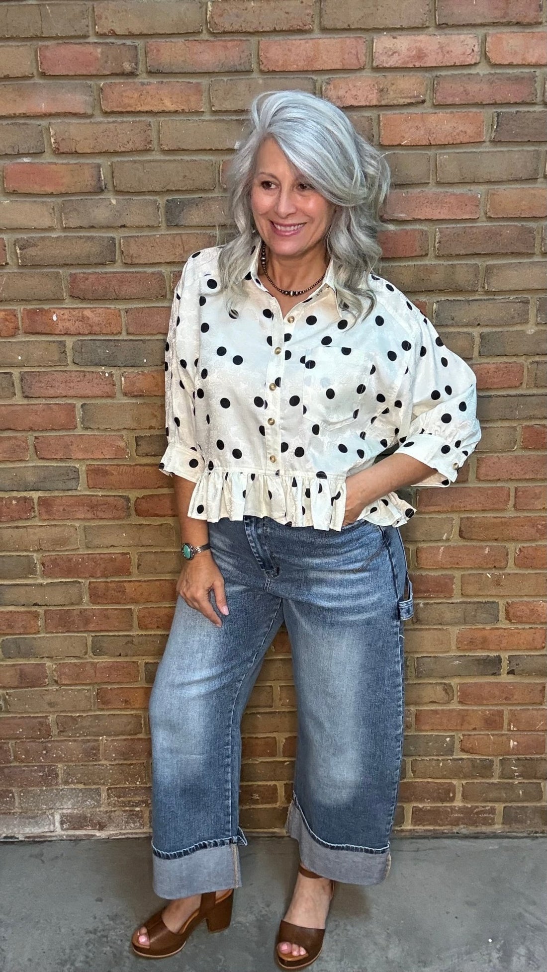 Woman wearing the Polka Dot Ruffle Hem Shirt and jeans against a brick wall.