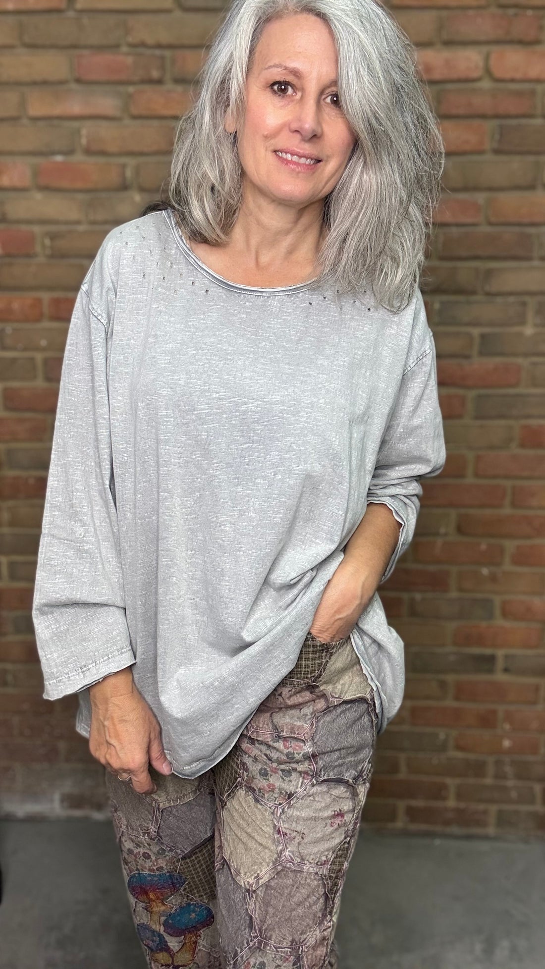 Woman wearing a gray oversized shirt and patterned pants standing against a brick wall.