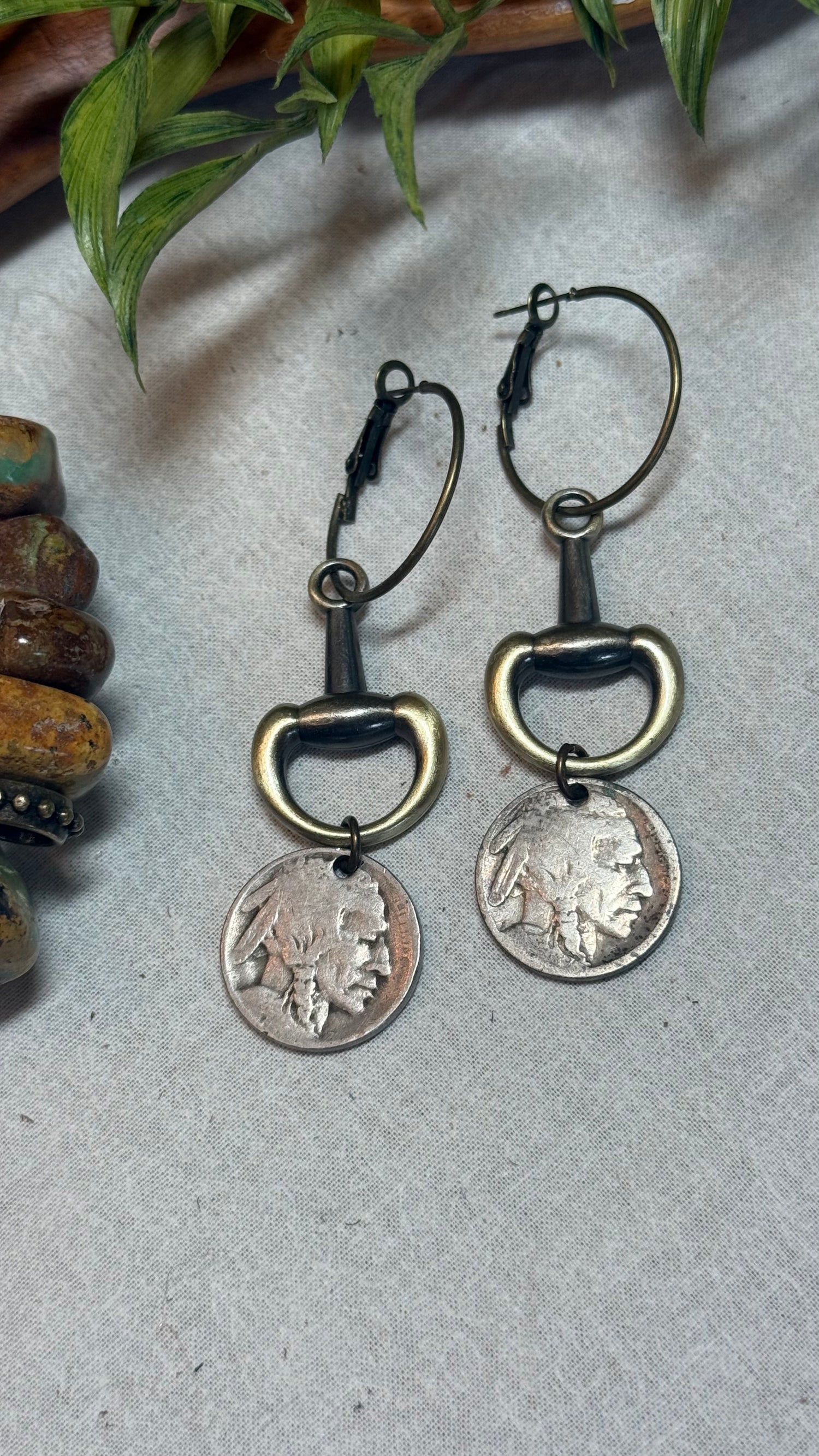 A pair of drop earrings featuring antique bronze horsebit rings and silver-toned US coins, displayed on a textured gray surface with decorative green leaves and wooden beads in the background.