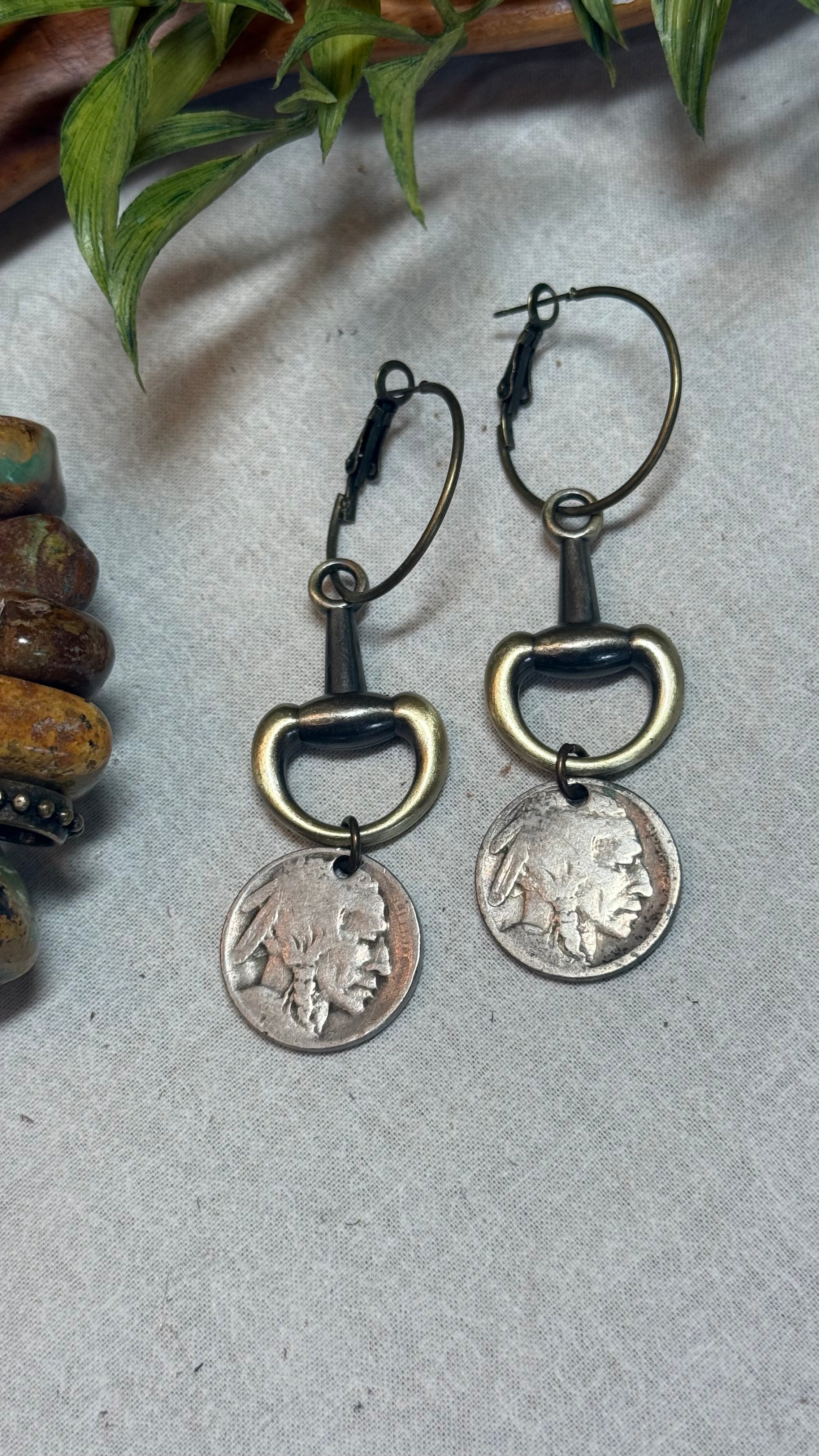 A pair of drop earrings featuring antique bronze horsebit rings and silver-toned US coins, displayed on a textured gray surface with decorative green leaves and wooden beads in the background.