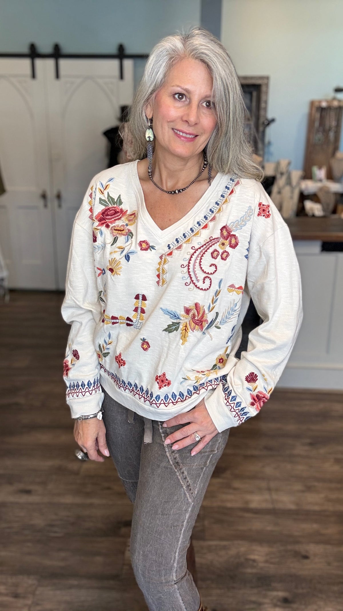 A woman with gray hair wearing a cream-colored long-sleeve V-neck sweater with colorful floral and paisley embroidery, paired with gray jeans, standing in a home interior setting.