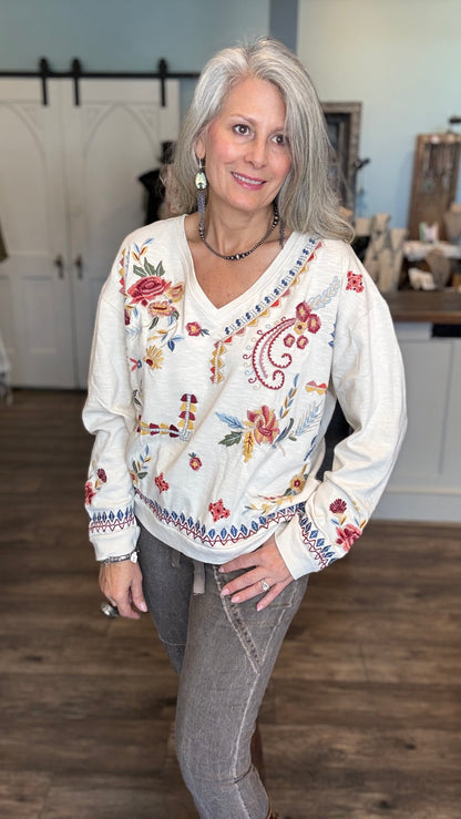 A woman with gray hair wearing a cream-colored long-sleeve V-neck sweater with colorful floral and paisley embroidery, paired with gray jeans, standing in a home interior setting.