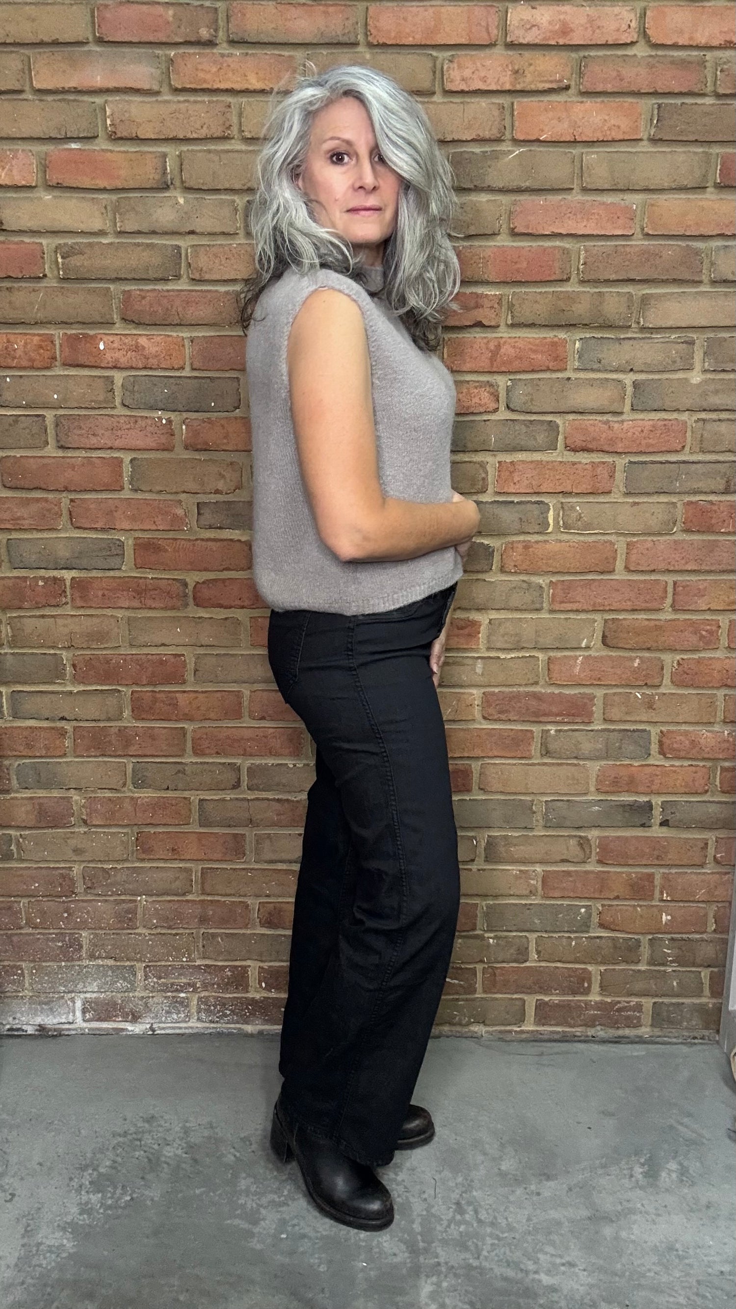 Woman wearing a gray sleeveless top and black pants against a brick wall.