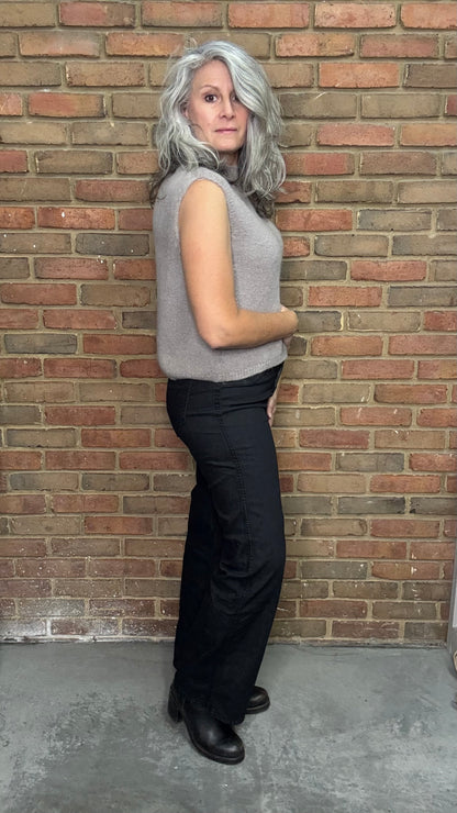 Woman wearing a gray sleeveless top and black pants against a brick wall.