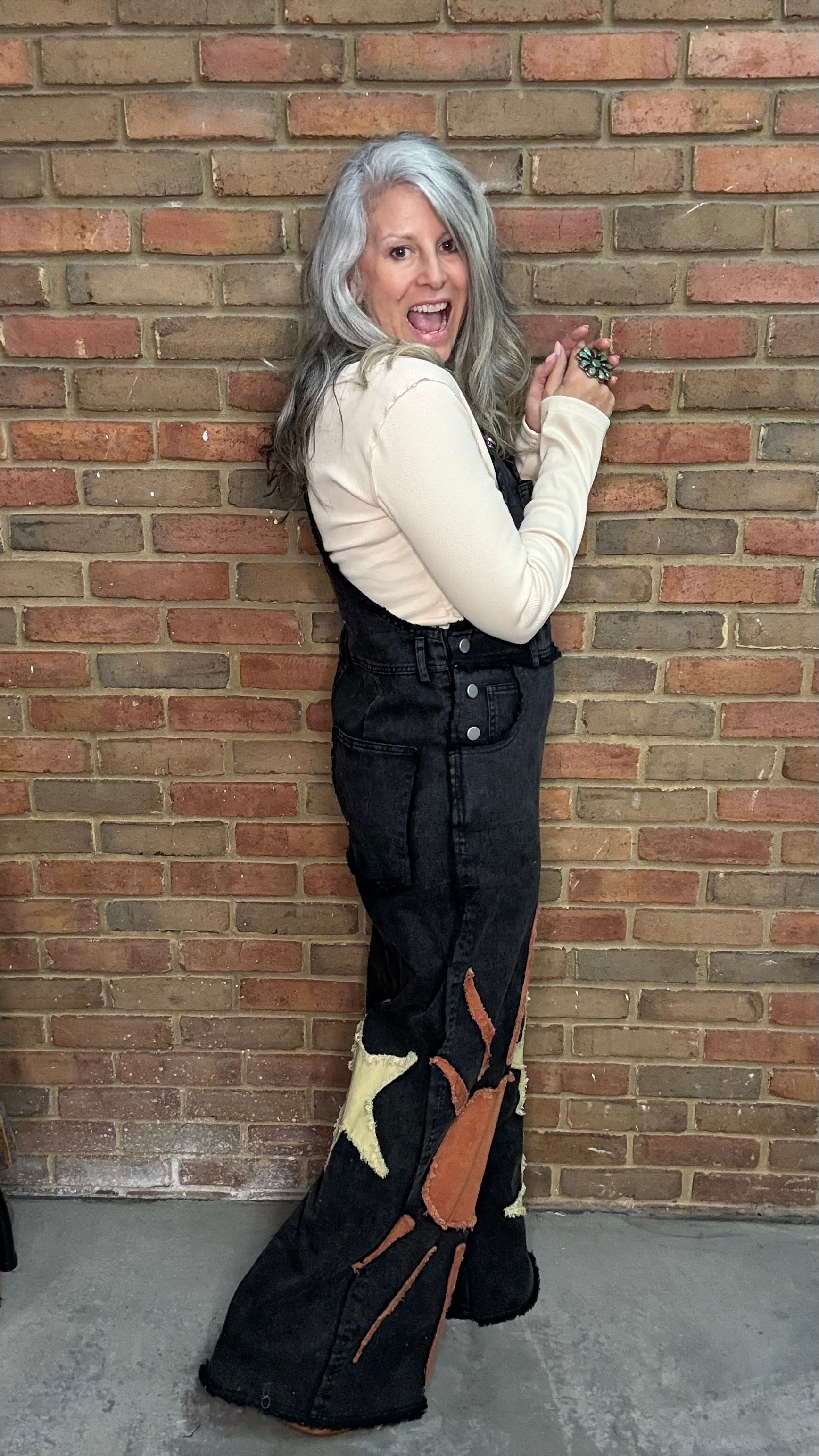 A woman with long gray hair poses in front of a brick wall wearing a cream long-sleeve top and black denim overall flare pants with a brown and beige abstract graphic print near the hem.